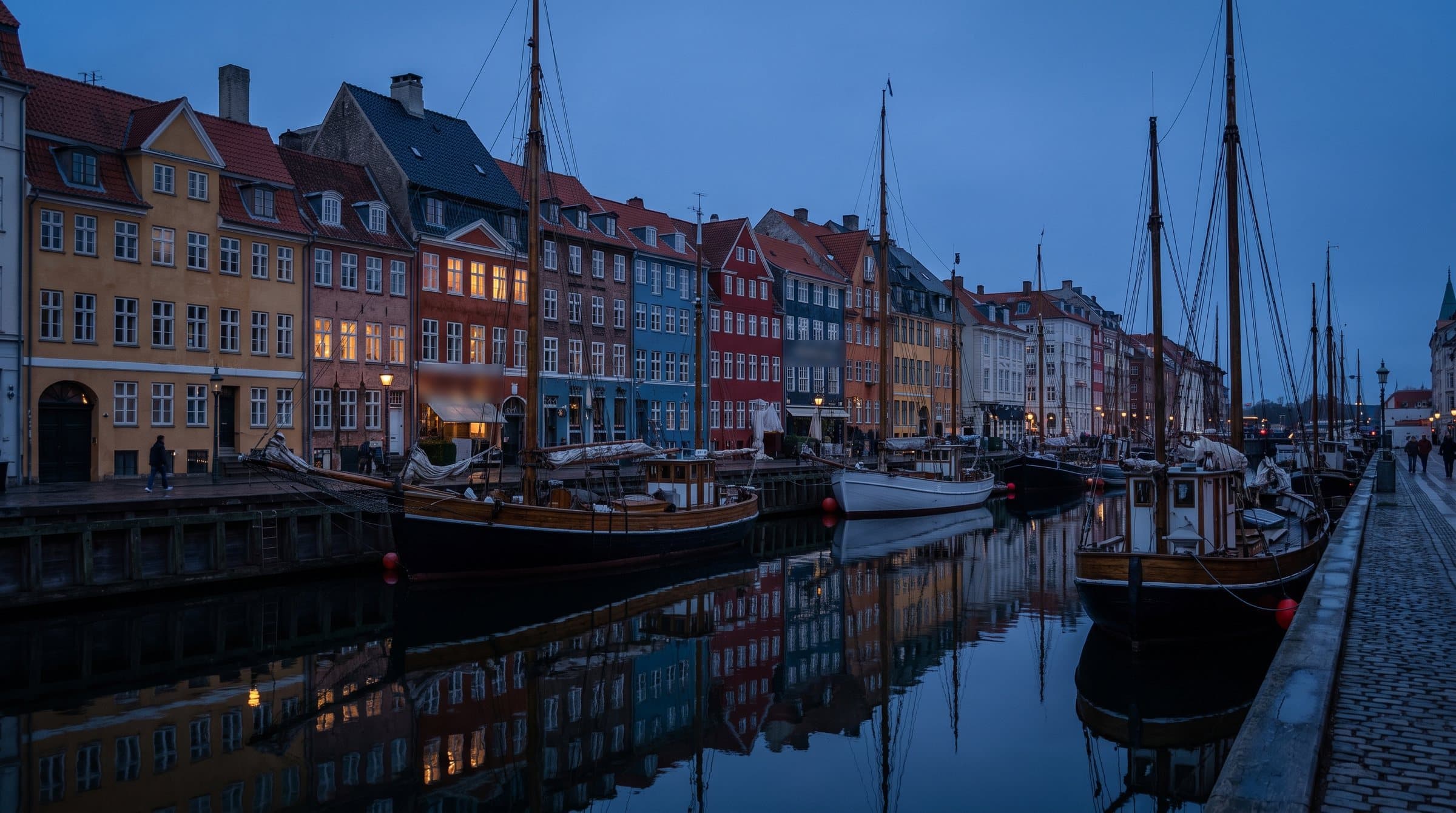 Nyhavn i København i den blå time: farverige gavlhuse med oplyste vinduer og gamle træskibe, der spejler sig i kanalen