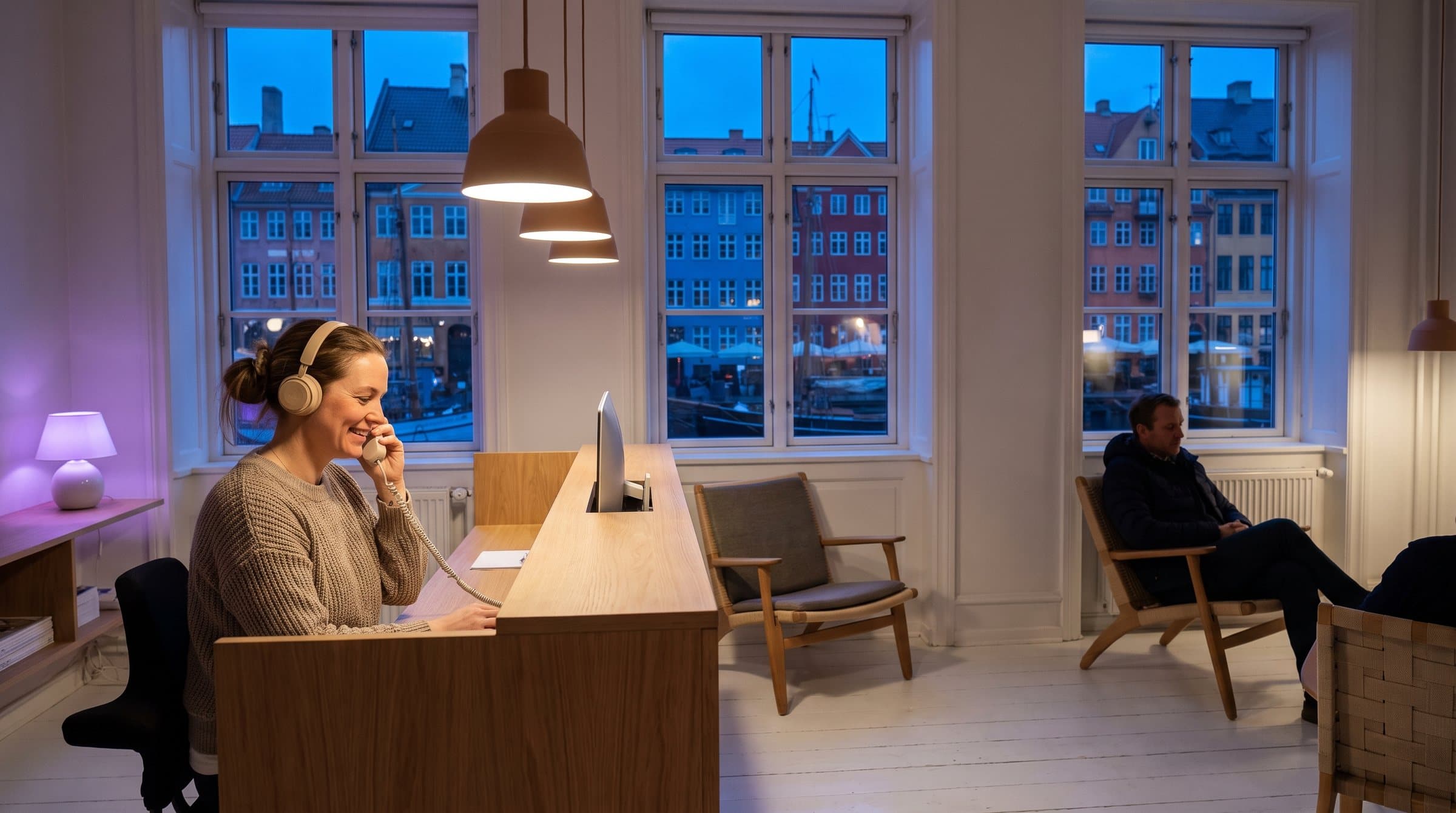Receptionist med headset tager telefonen i receptionen på en tandklinik i et gammelt københavnerhus med udsigt til de farverige huse i Nyhavn i den blå time